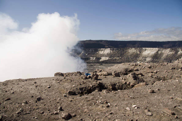 VolcanoWatch082709 A lone blue plastic bucket sampler braves the crater’s edge near the Overlook vent with HVO on the horizon. (Photo courtesy of Hawaiian Volcano Observatory)