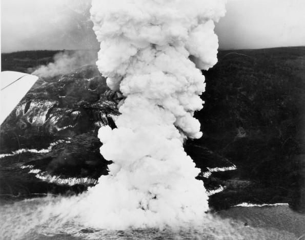During the Mauna Loa 1950 eruption, three large, fast-moving lava flows advanced down the west flank of the volcano, each crossing the main highway before reaching the ocean. The Ka‘apuna flow, shown here, traveled from the Southwest Rift Zone vent (7,800 ft elevation) to the coast in just 17 hours, creating a huge steam plume as lava flowed into the sea. The glowing edges and fast-moving channels of the 10- to 25-ft thick ‘a‘ā flow appear white in this June 2, 1950, aerial image. Photo courtesy of Air National Guard, 199th Fighter Squadron.