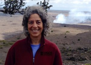 Tamar Elias at the rim of Kīlauea's summit caldera, with Halemaʻumaʻu Crater in background (fumes are from the lava lake within the crater). Photo courtesy U.S. Geological Survey Hawaiian Volcano Observatory.