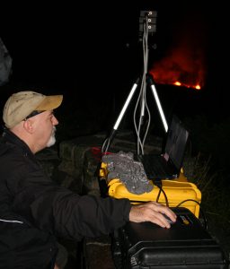 A U.S. Geological Survey scientist used a custom-built, high-speed camera to remotely measure the temperature of spattering lava on the surface of Kīlauea Volcano’s summit lava lake (background). This particular experiment, conducted from the Jaggar Museum overlook in Hawaiʻi Volcanoes National Park on December 10, 2016, was part of a NASA-funded research project. USGS photo.
