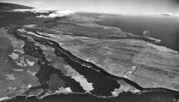 Most of the lava flow (dark black) produced by the April 1868 Mauna Loa eruption can be seen in this aerial photo on the west (left) side of the prominent fault scarp, known informally as the Kahuku pali (formal names are Pali o Mamalu for the upper half and Pali‘okūlani for the lower half of the scarp). The large littoral cone that formed during the eruption, now named Pu‘uhou, is visible on the lower left coastline. Kalae (South Point) is not visible, but is to the right of the photo. The summit areas of Mauna Loa and Kīlauea volcanoes can be seen in the distance. Aerial photo: U.S. Geological Survey, 1954.